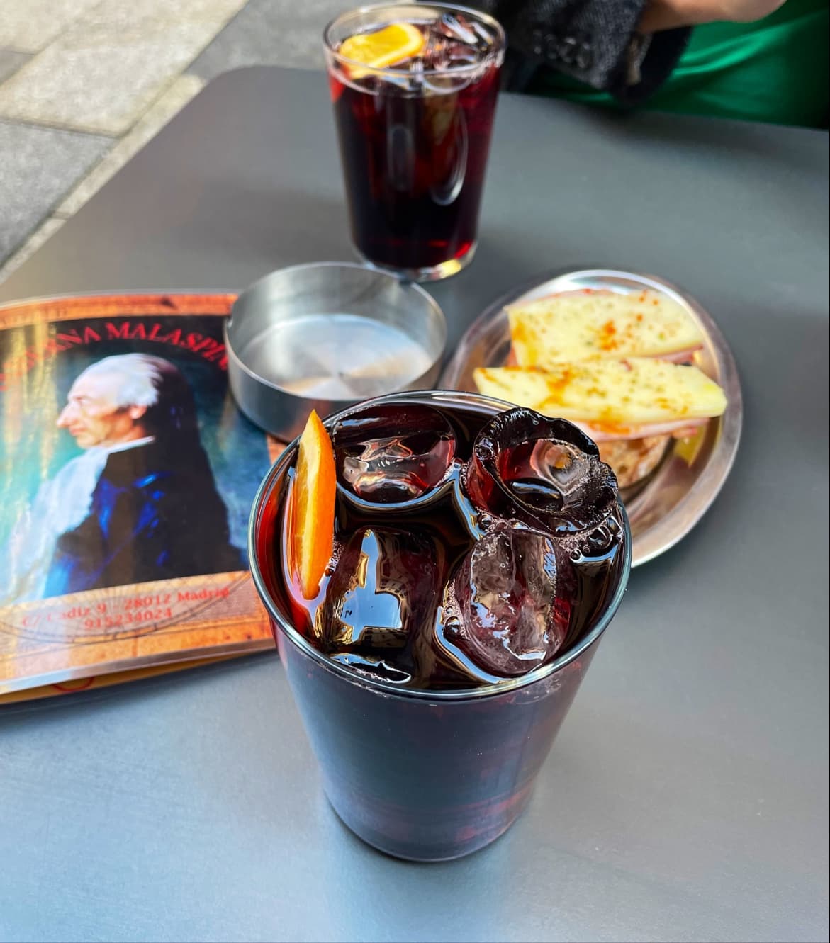 Dos bebidas oscuras con naranja y una tapa en la mesa exterior de Malaspina